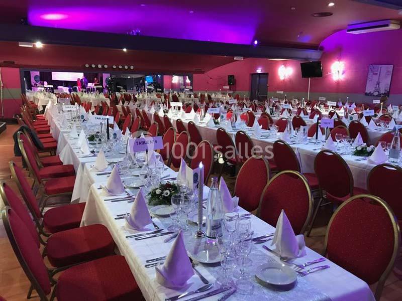 Salle de réception avec de longues tables dressées, nappes blanches, serviettes pliées et chaises rouges.
