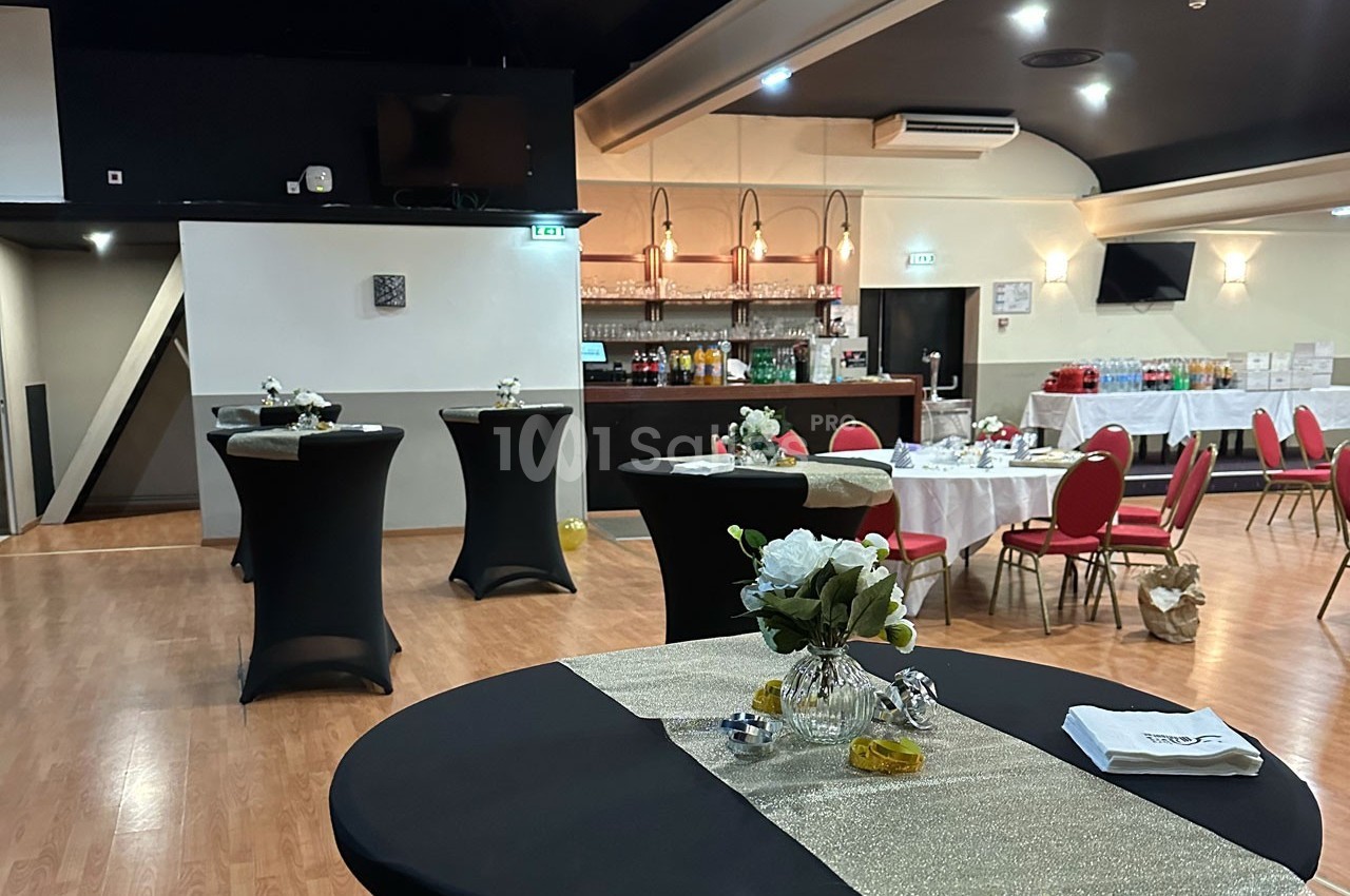 Salle de réception avec tables hautes décorées, bar en arrière-plan et tables rondes dressées pour un événement.