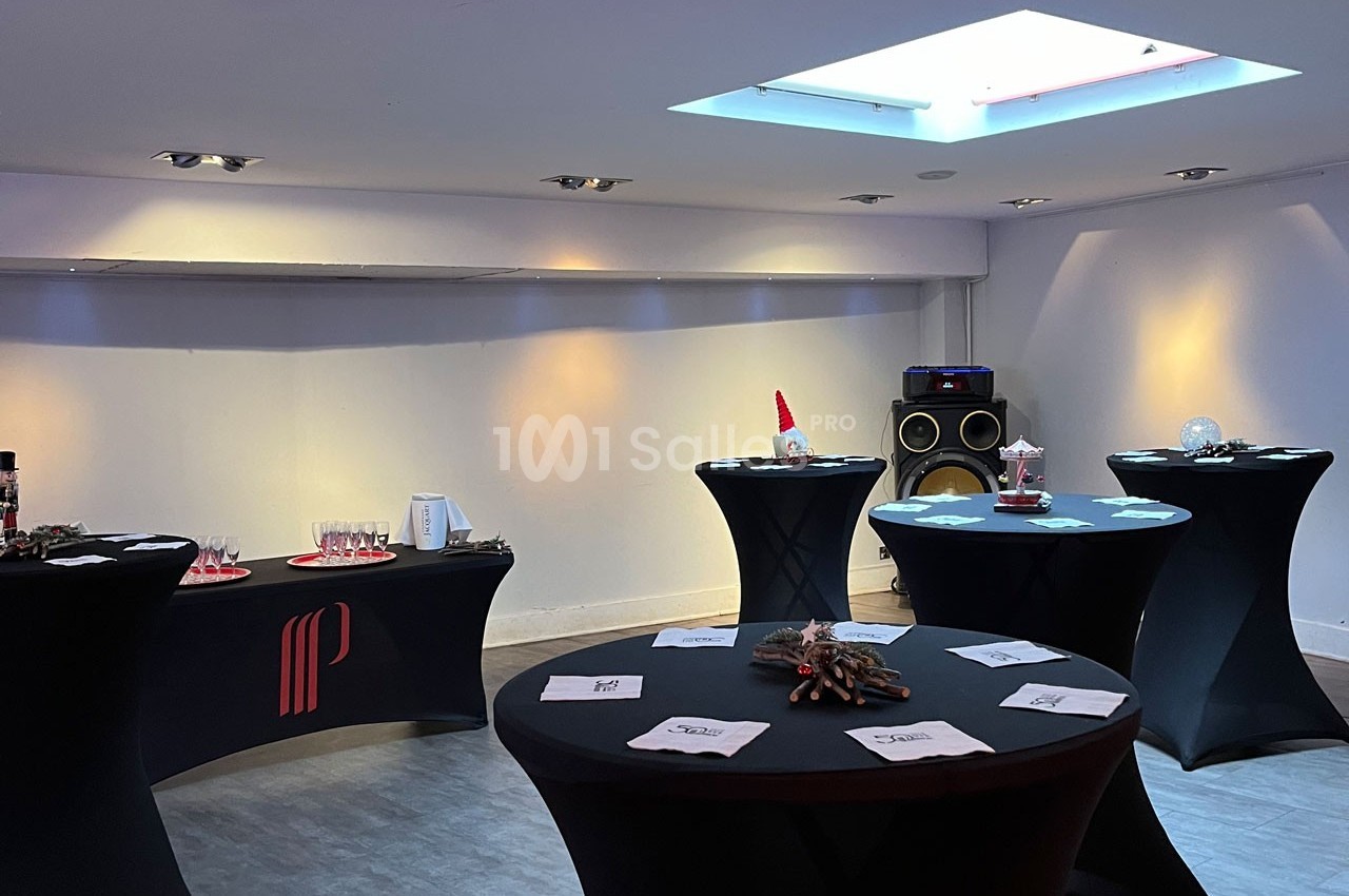 Salle lumineuse avec tables hautes noires dressées pour un événement, éclairage tamisé et enceinte visible au fond.