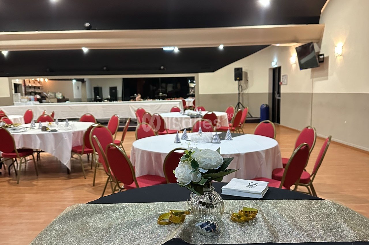 Salle de réception avec tables rondes dressées, chaises rouges et décoration florale sur une table au premier plan.