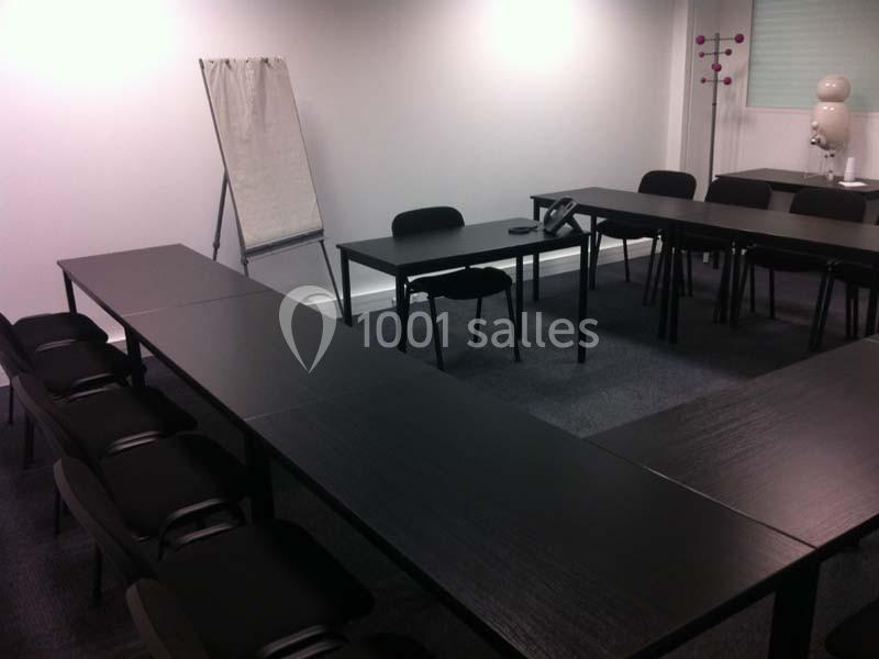 Salle de réunion vide avec des tables noires disposées en U, un chevalet avec feuille et des chaises noires.
