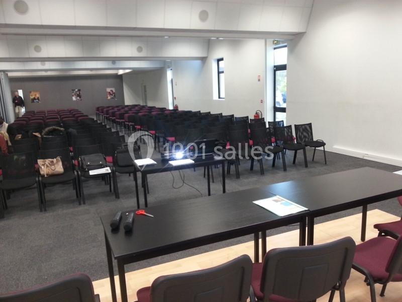 Salle de conférence vide avec des rangées de chaises, une table équipée d'un projecteur et des documents posés dessus.