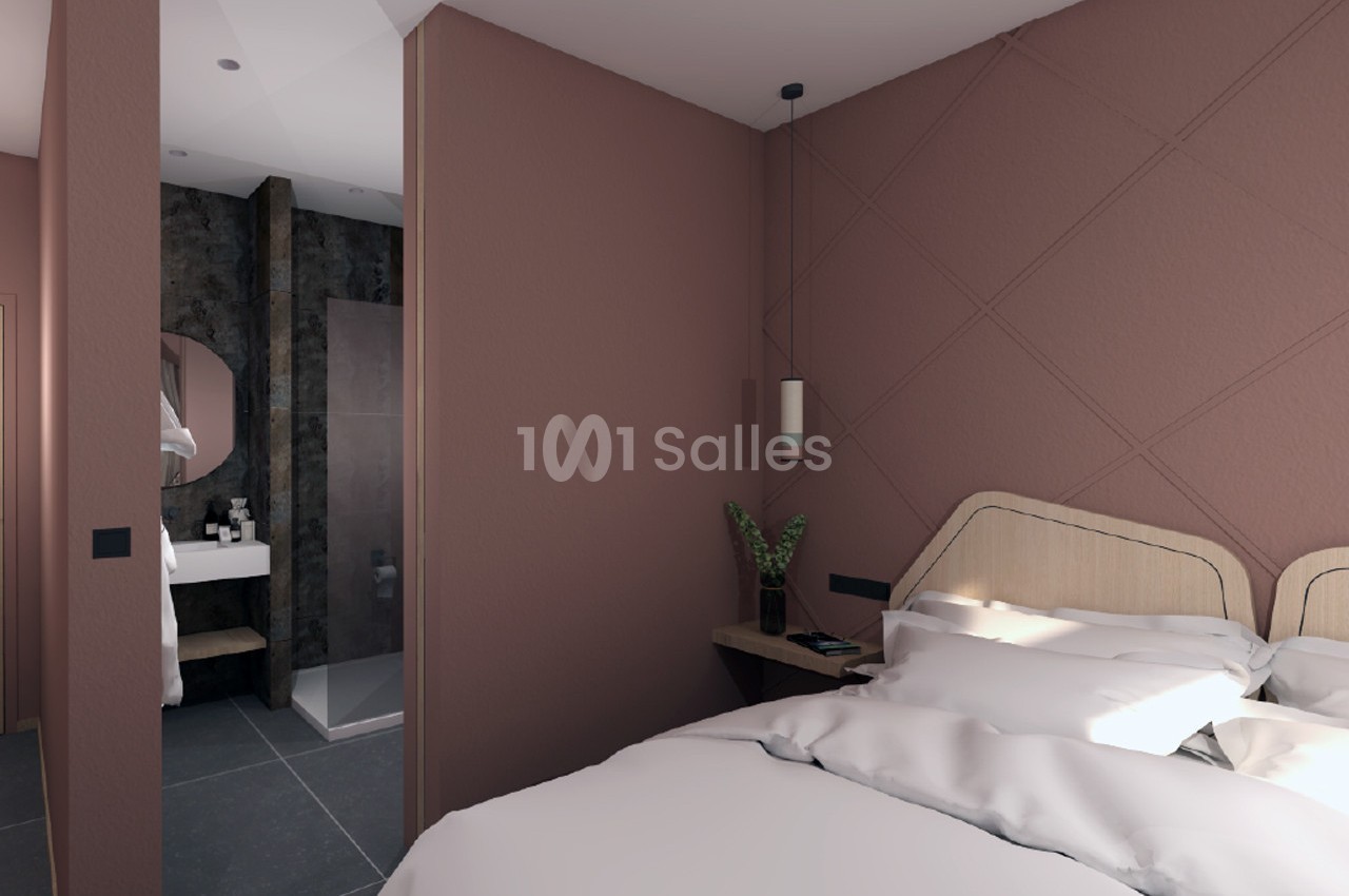 Chambre moderne avec lit double, murs roses, suspension lumineuse et vue partielle sur une salle de bain attenante.
