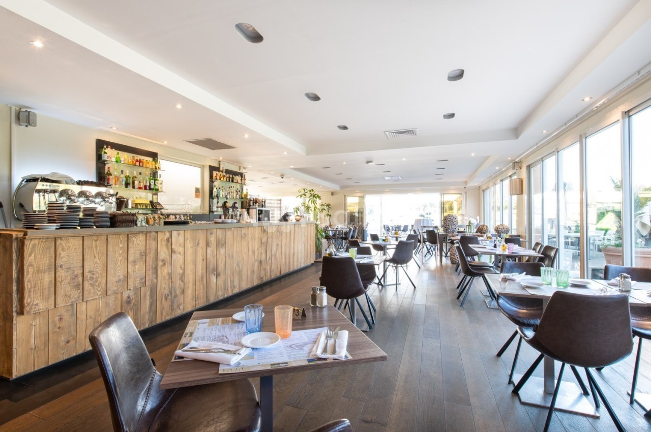 Salle de restaurant lumineuse avec un bar en bois, des tables dressées et de grandes baies vitrées donnant sur l'extérieur.