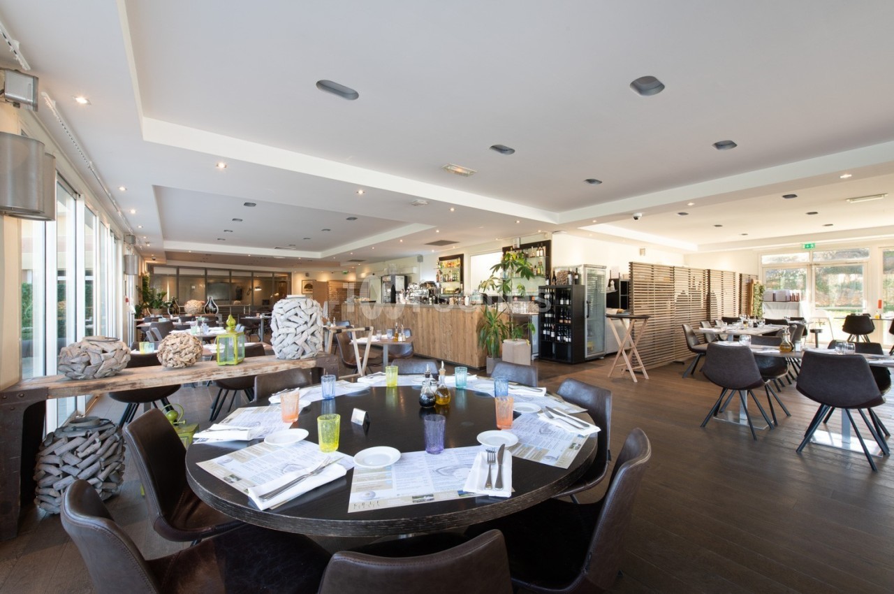 Salle de restaurant lumineuse avec tables dressées, chaises noires et grandes baies vitrées donnant sur l'extérieur.