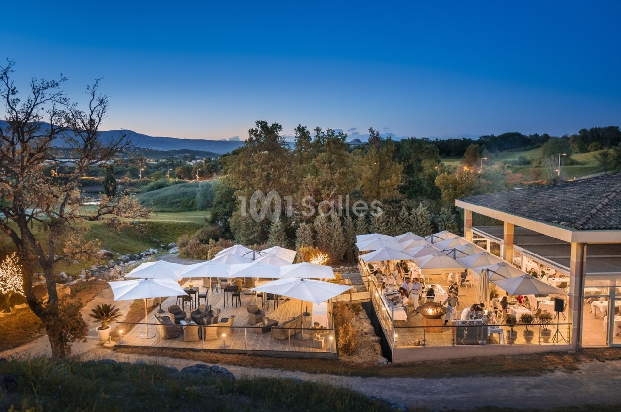Terrasse d'un restaurant éclairée en soirée, avec des tables sous des parasols et vue sur un paysage naturel.