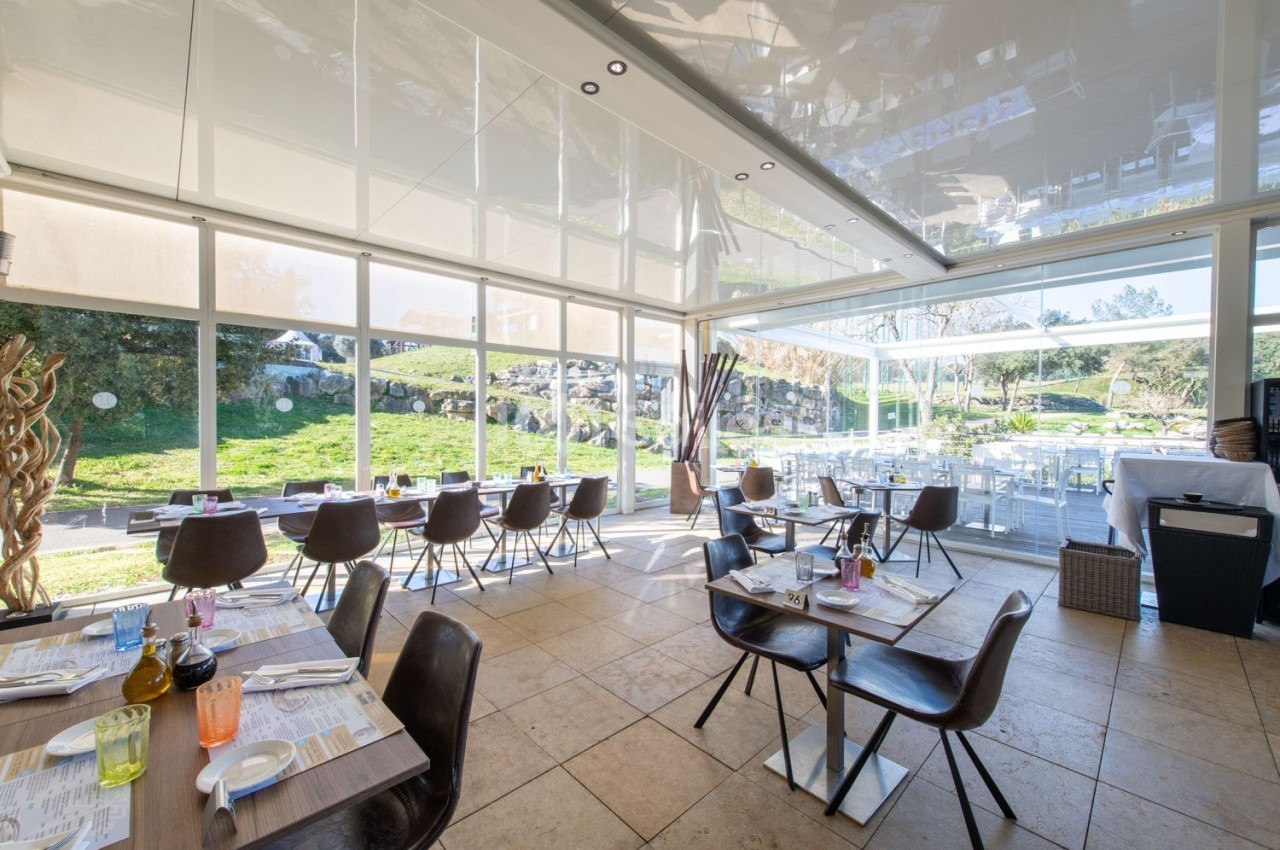 Salle de restaurant lumineuse avec de grandes baies vitrées donnant sur un jardin verdoyant.