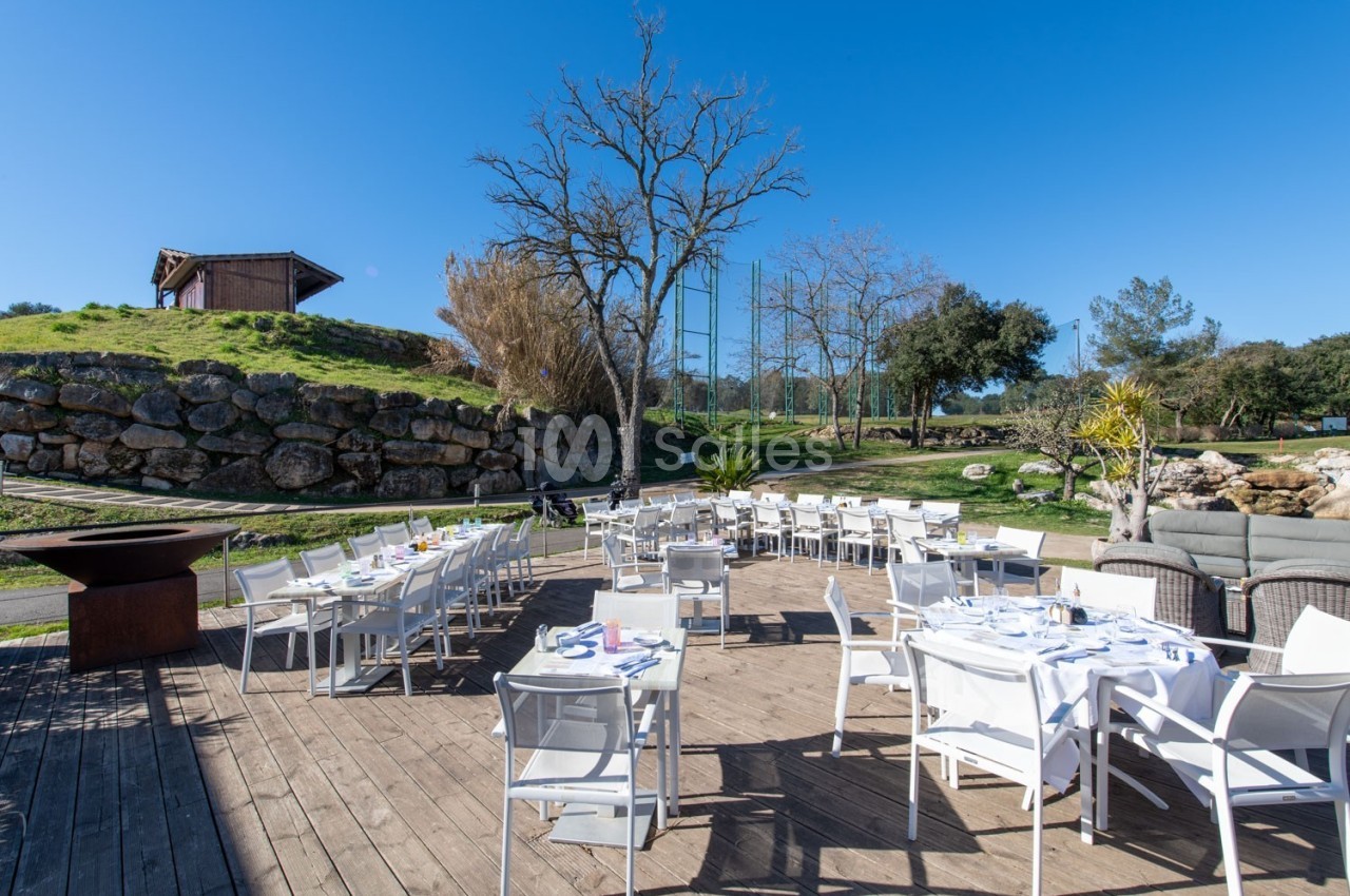 Terrasse en bois avec des tables et chaises blanches dressées, entourée de verdure et d'un ciel dégagé.