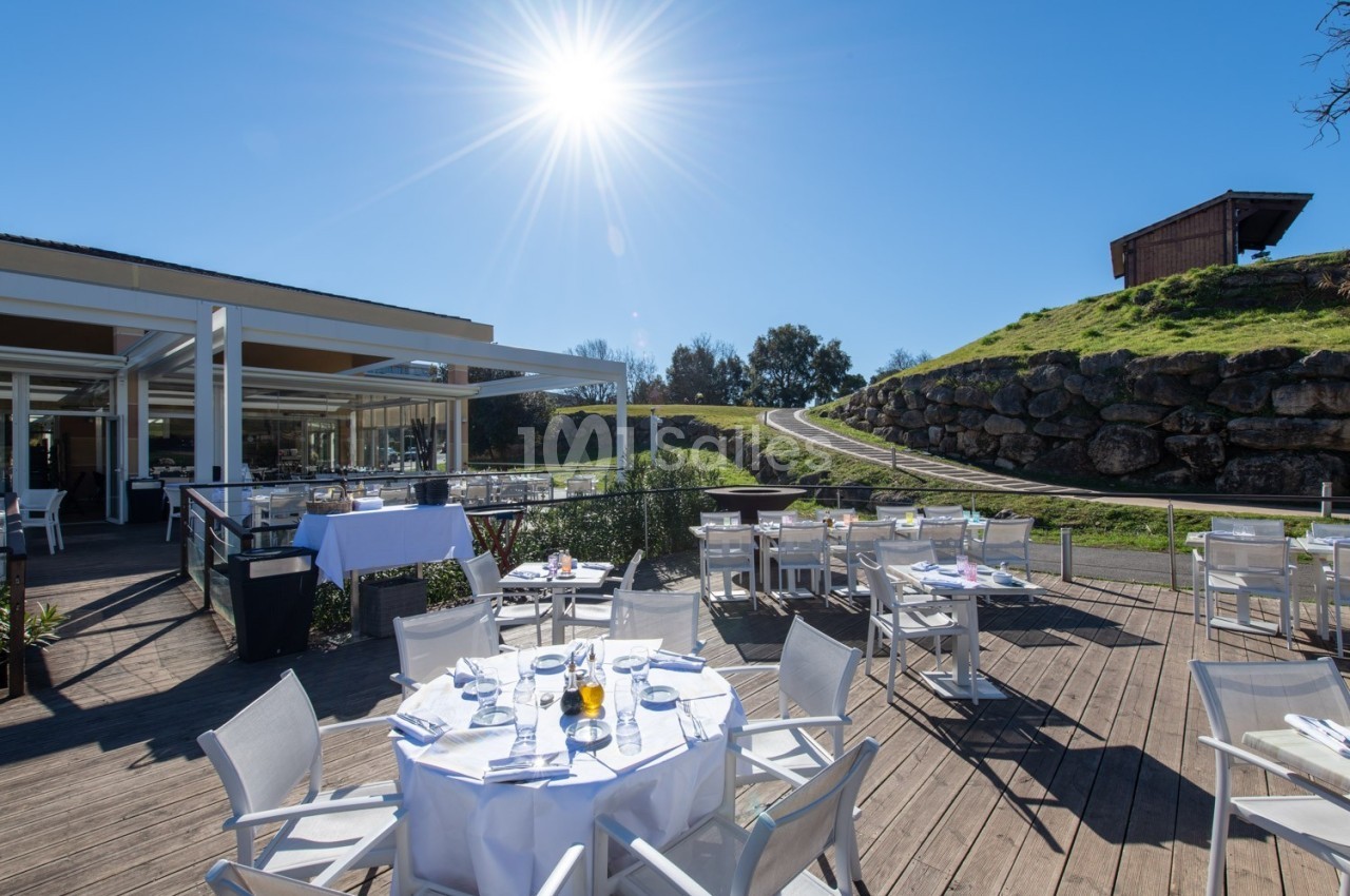 Terrasse ensoleillée avec tables dressées, entourée de verdure et d'une petite colline sous un ciel bleu.