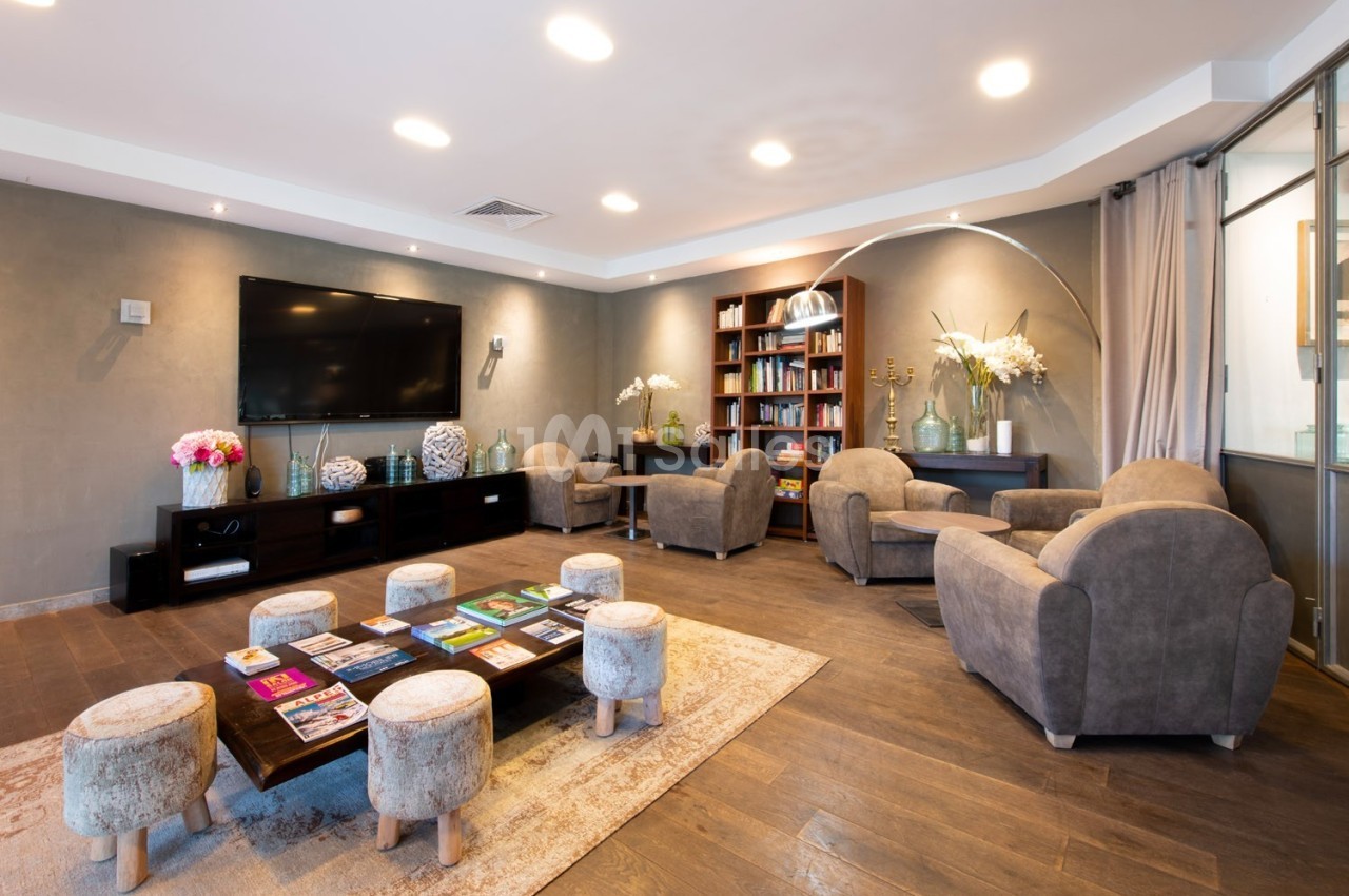 Salon moderne avec fauteuils, table basse, étagères remplies de livres, écran mural et éclairage chaleureux.
