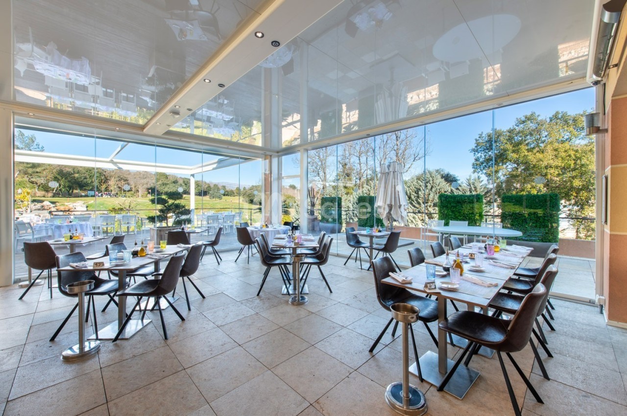 Salle de restaurant lumineuse avec grandes baies vitrées donnant sur un paysage verdoyant et des tables dressées.