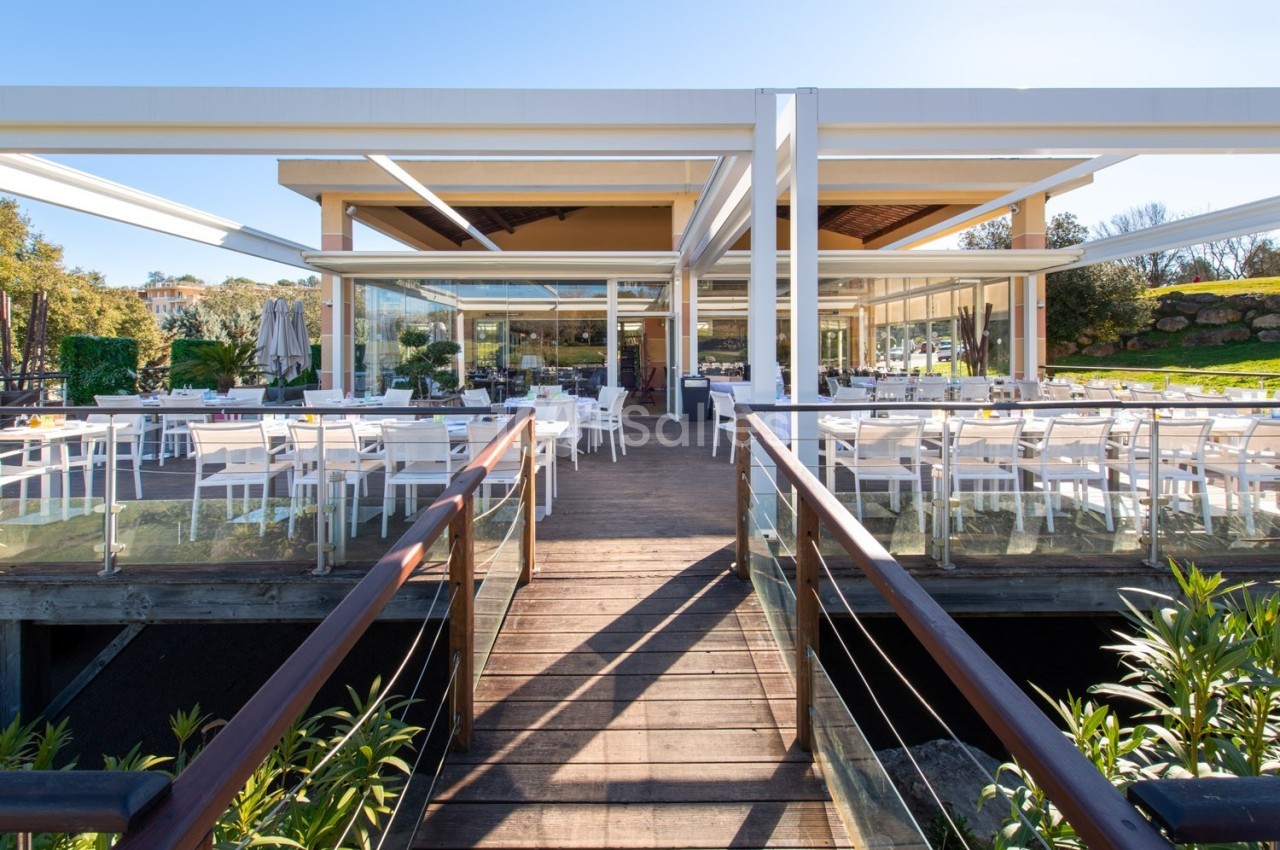 Terrasse d'un restaurant moderne avec tables et chaises blanches, entourée de verdure et éclairée par la lumière du jour.