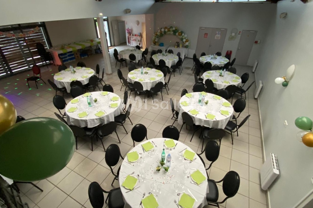 Salle décorée avec des tables rondes dressées, nappes blanches, serviettes vertes et ballons pour un événement festif.