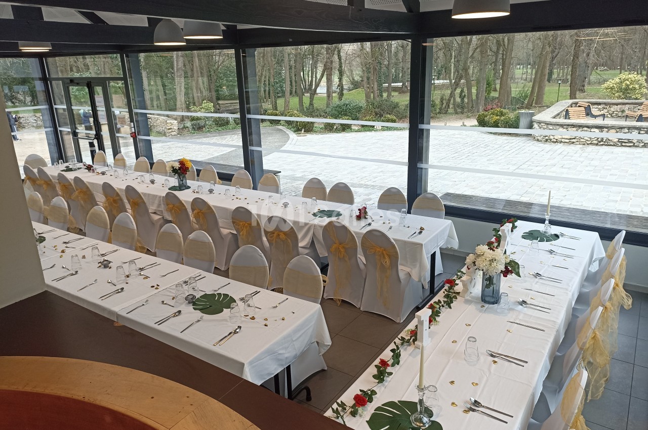 Salle de réception avec deux longues tables dressées, décorées de fleurs et nappes blanches, vue sur un jardin extérieur.