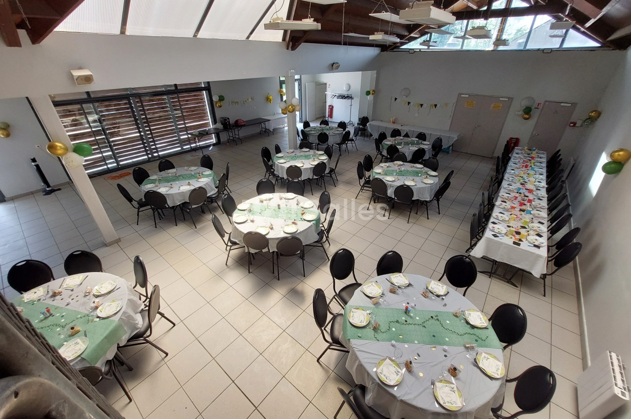 Salle décorée avec des tables rondes et une longue table, préparées pour un événement avec nappes vertes et vaisselle.