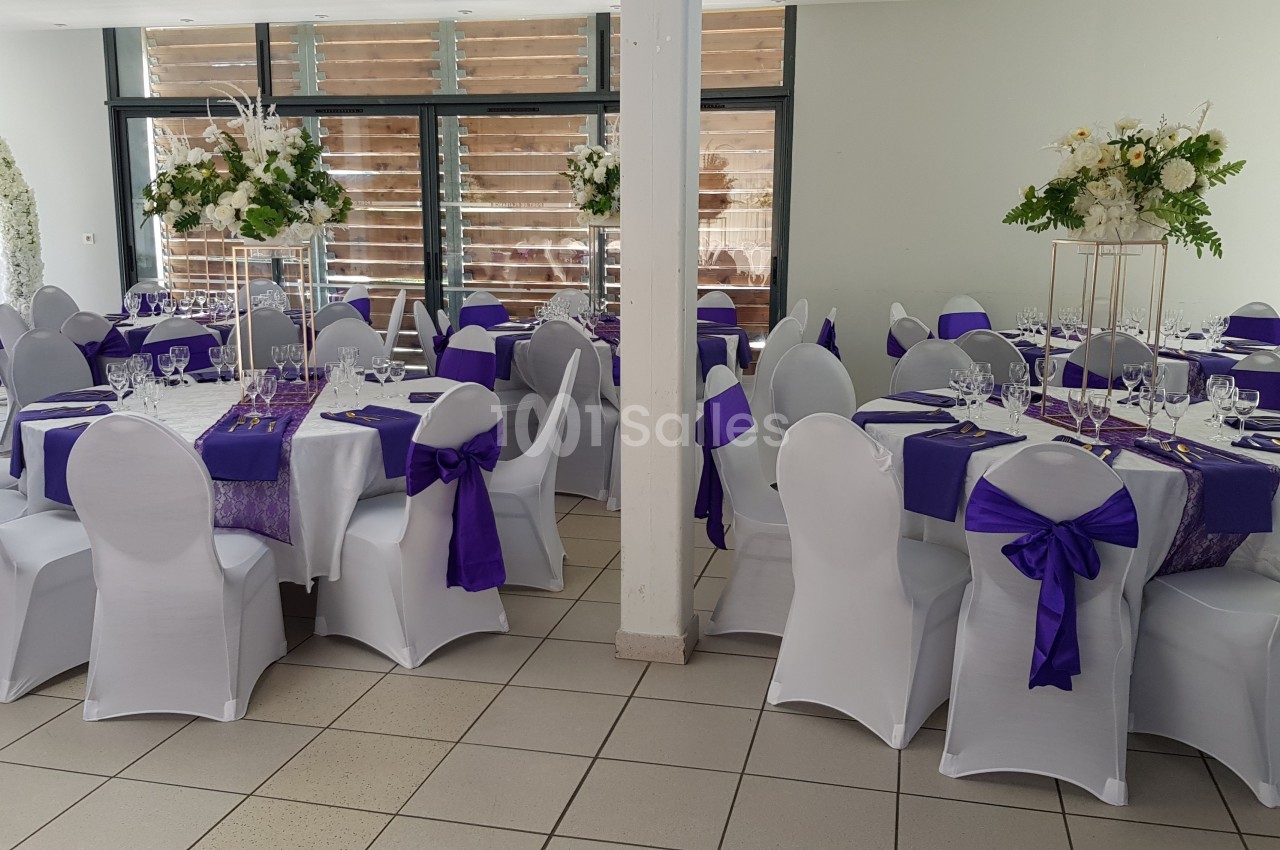 Salle décorée pour un événement avec des tables rondes dressées, nappes blanches et chemins de table violets.