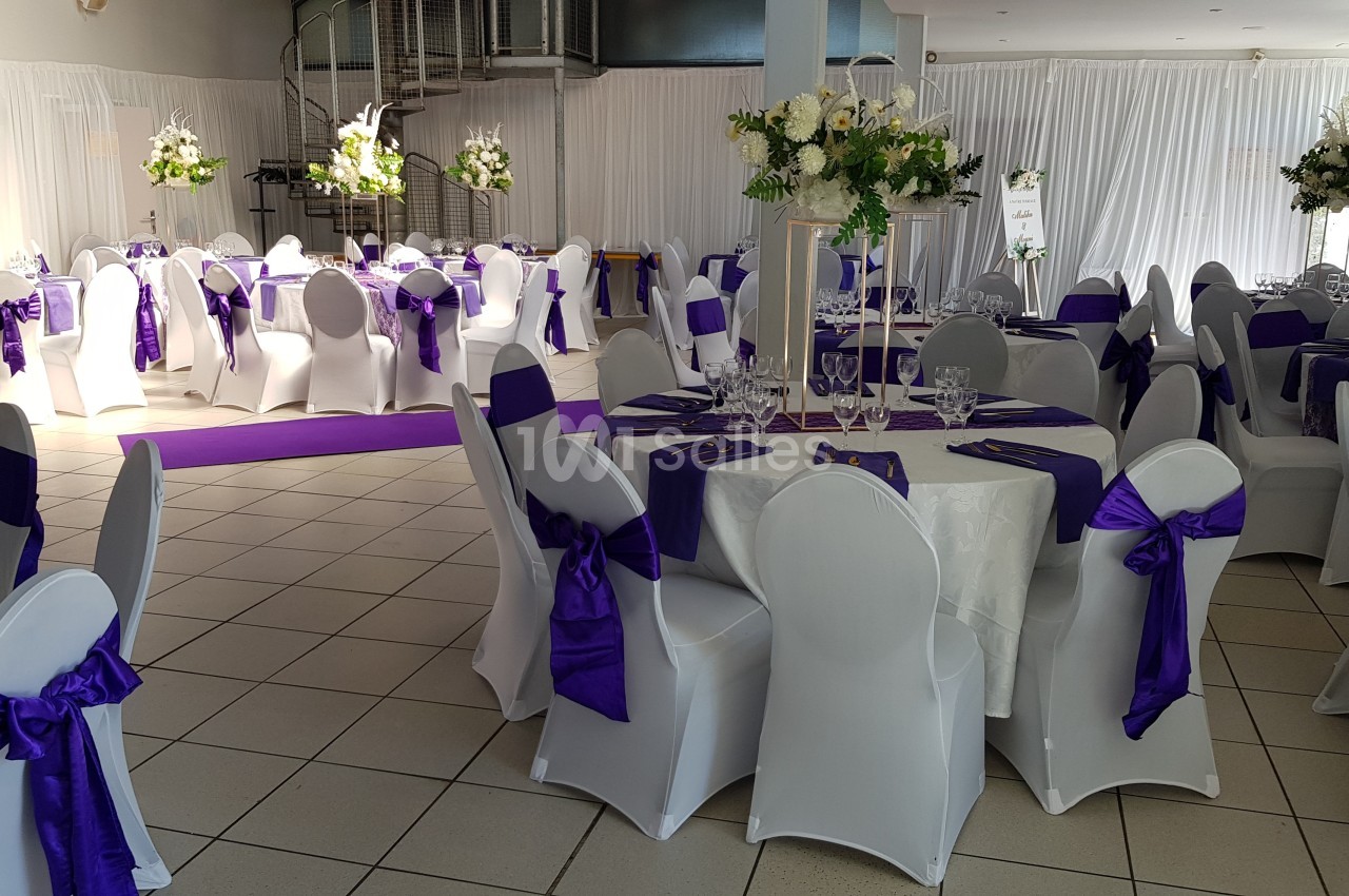 Salle de réception décorée avec des nappes blanches, des chaises ornées de nœuds violets et des arrangements floraux.