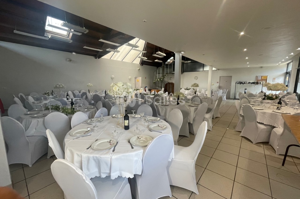 Salle de réception lumineuse avec des tables rondes dressées, nappes blanches et chaises couvertes.