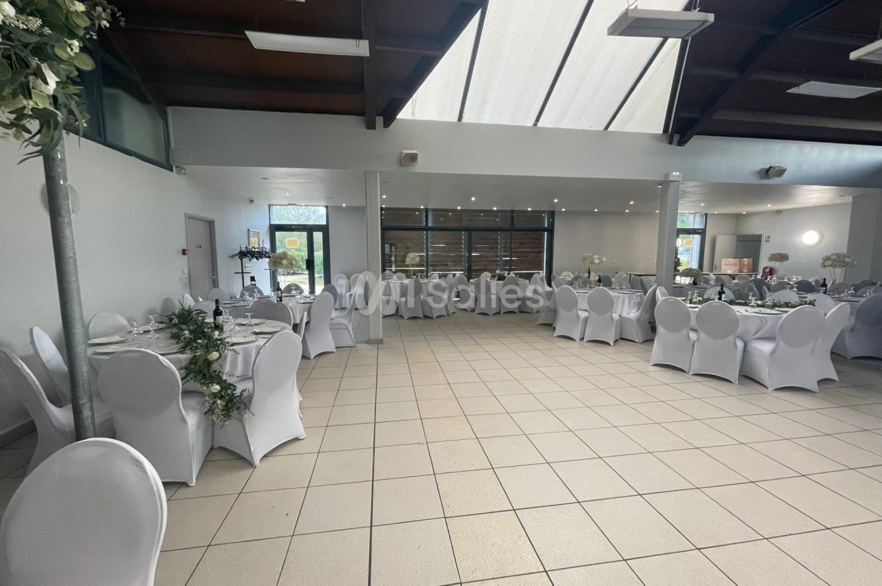 Salle de réception lumineuse avec tables rondes dressées, chaises blanches et décoration florale.
