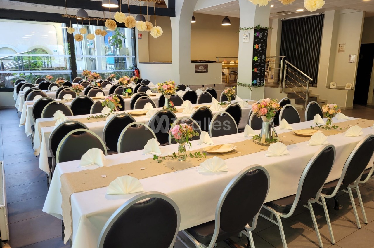Salle décorée pour un événement, avec des tables alignées, nappes blanches, fleurs et pliages de serviettes.