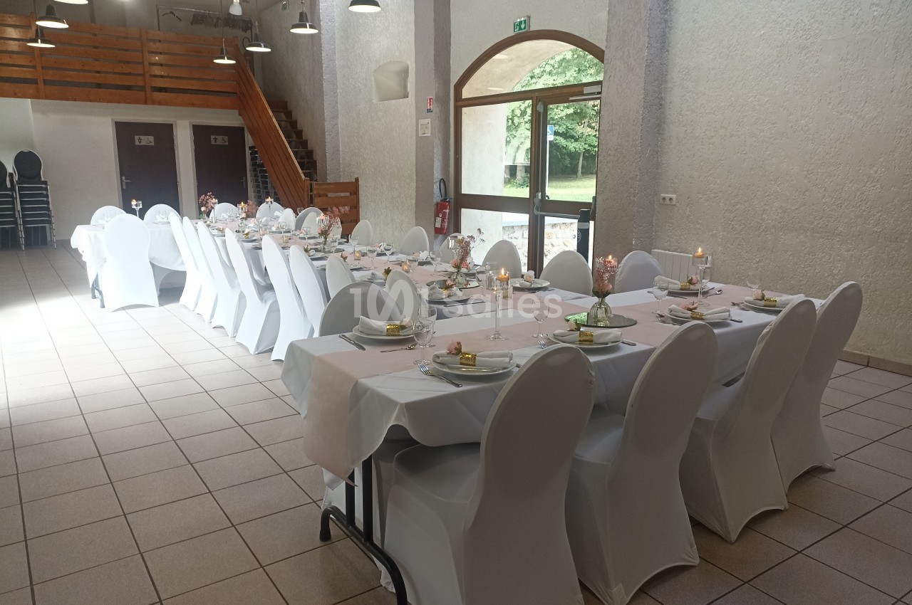 Salle décorée pour un repas, avec tables alignées, nappes blanches, chaises houssées et lumière naturelle entrant par une…