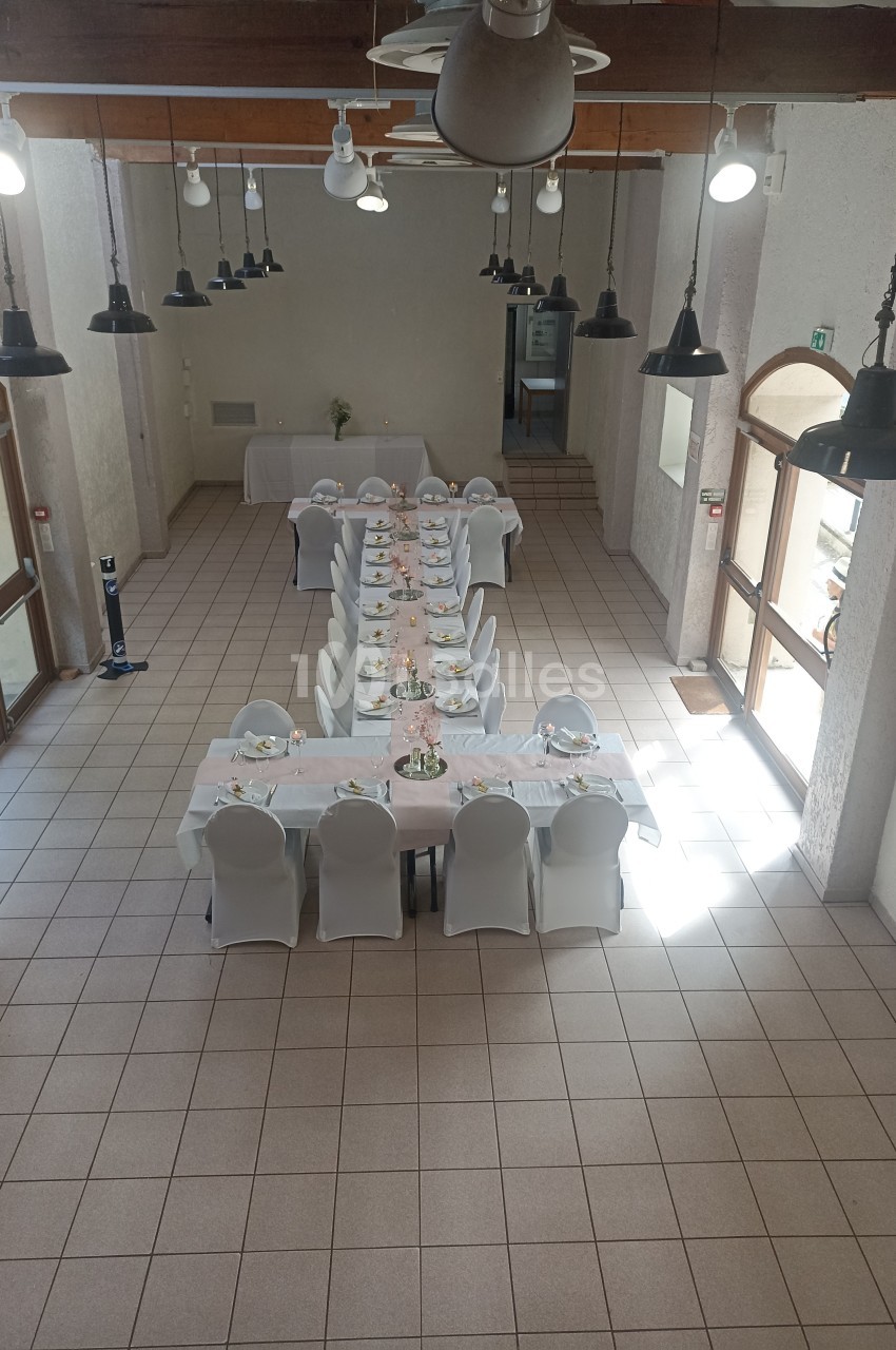 Salle lumineuse avec une grande table dressée pour un repas, entourée de chaises blanches, sous des lampes suspendues.