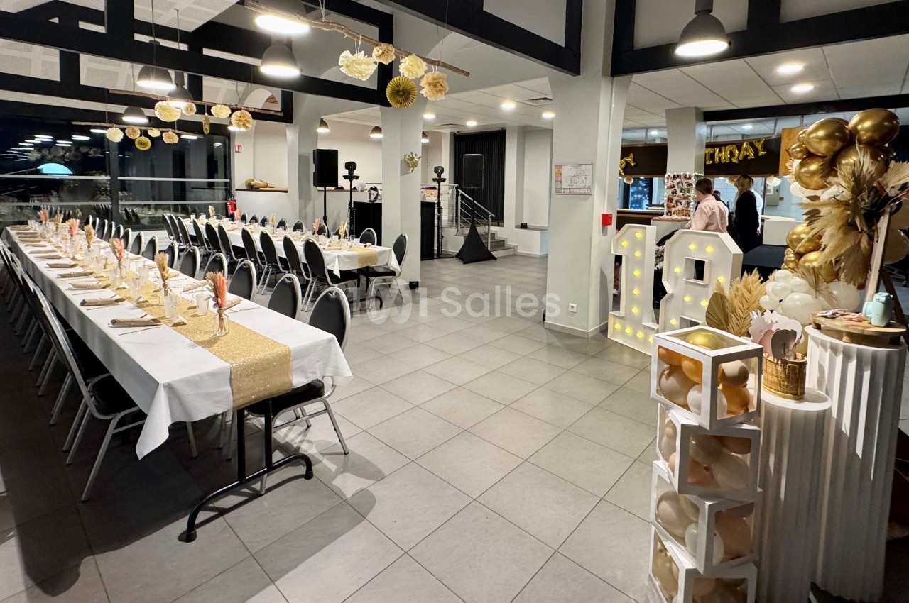 Salle décorée pour une fête, avec longues tables dressées, ballons dorés et blancs, et éléments lumineux.