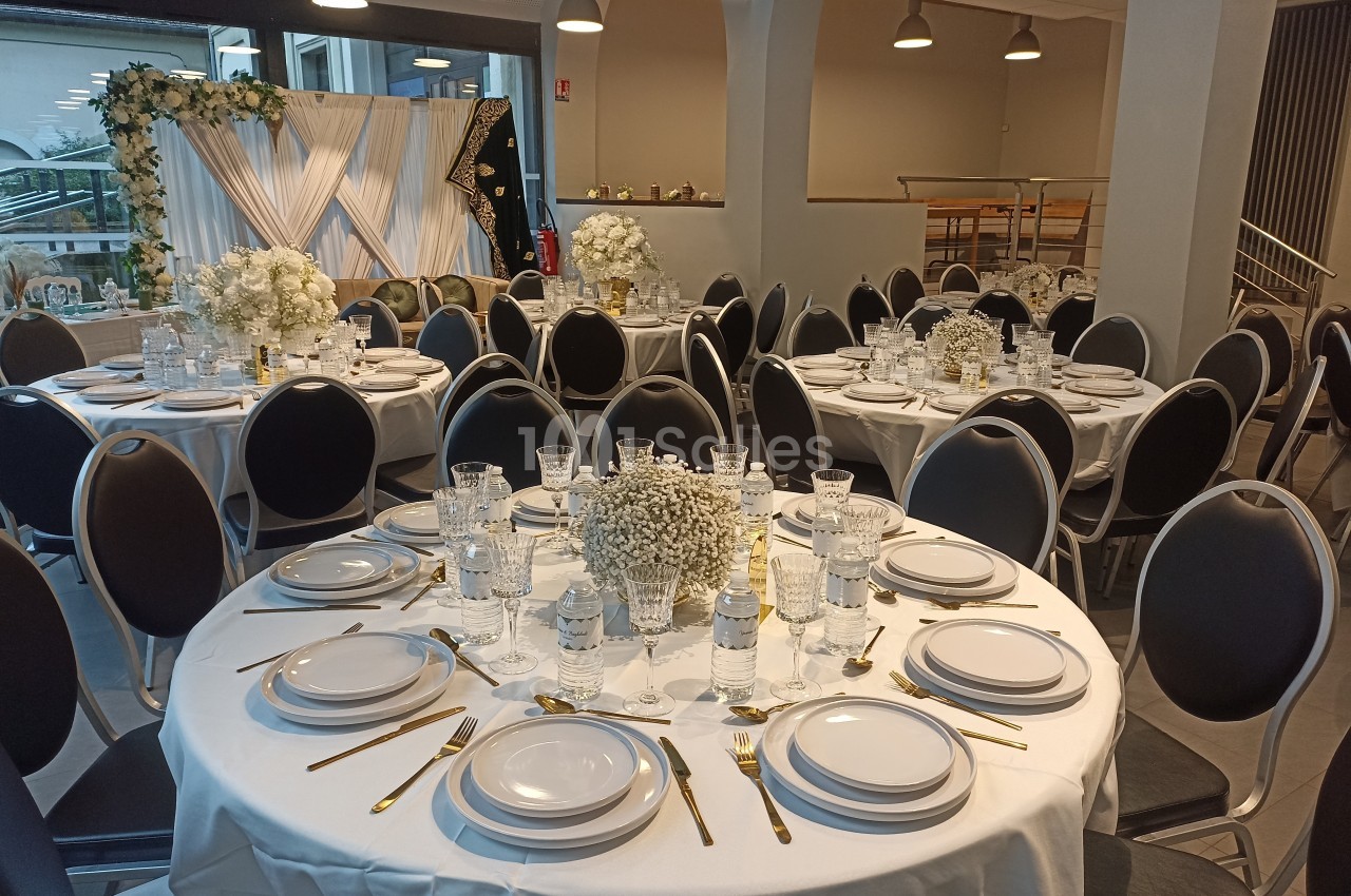 Salle de réception avec tables rondes dressées, nappes blanches, vaisselle élégante et décorations florales blanches.