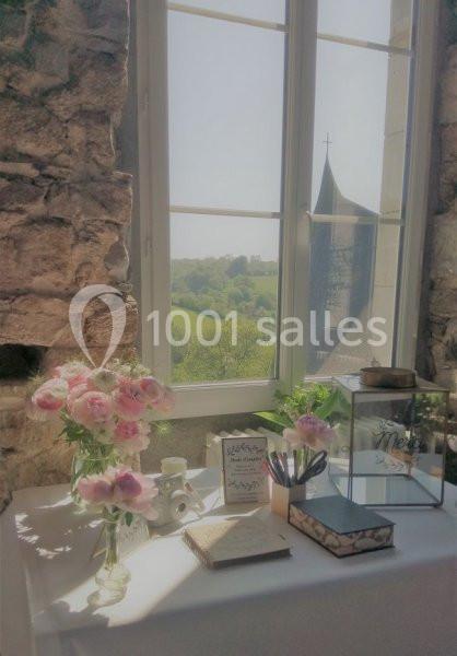 Table décorée avec des fleurs roses, papeterie et accessoires, près d'une fenêtre offrant une vue sur la campagne et une…