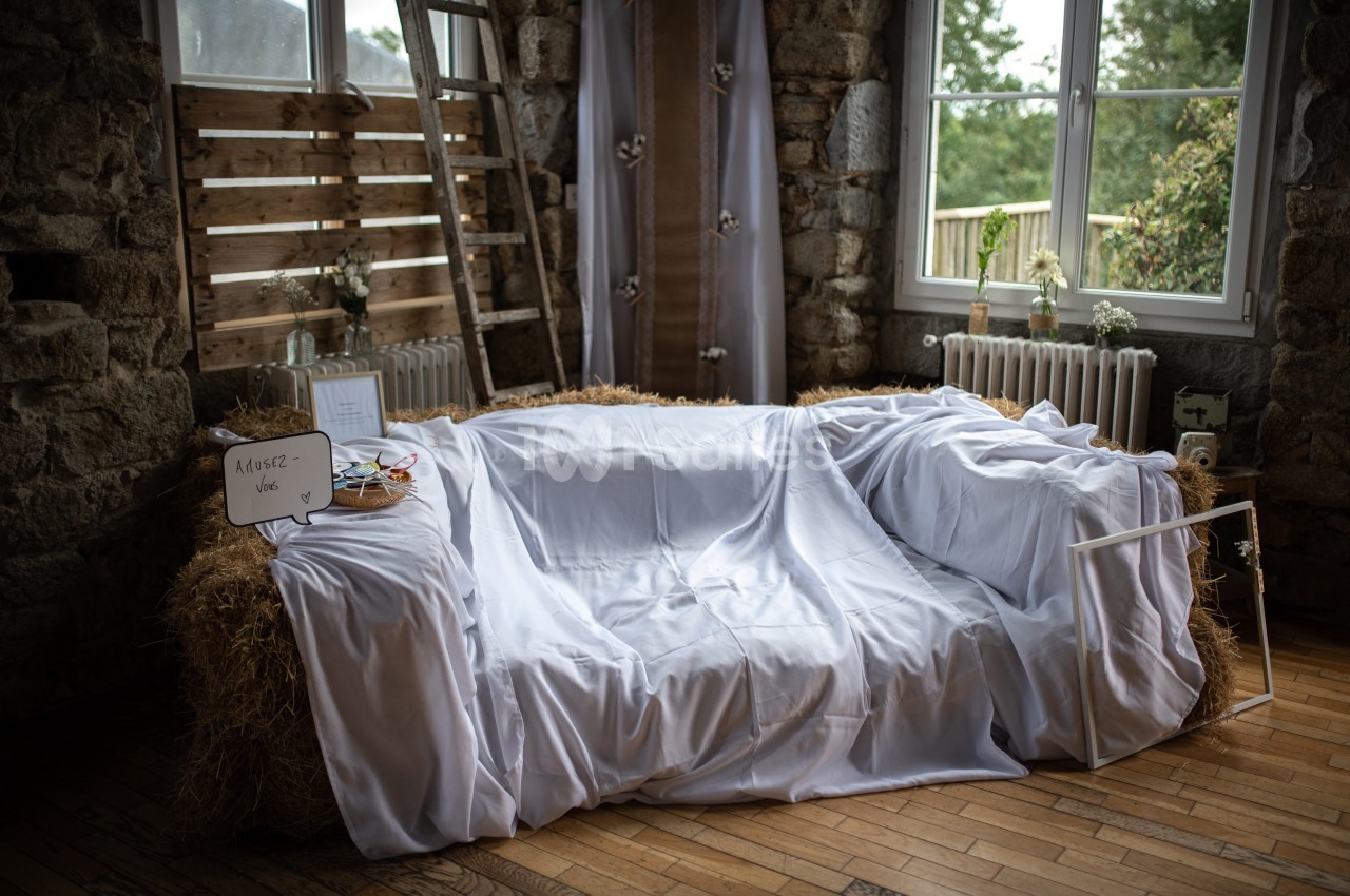Canapé improvisé en bottes de paille recouvertes de draps blancs, dans une pièce rustique avec fenêtres et décoration…