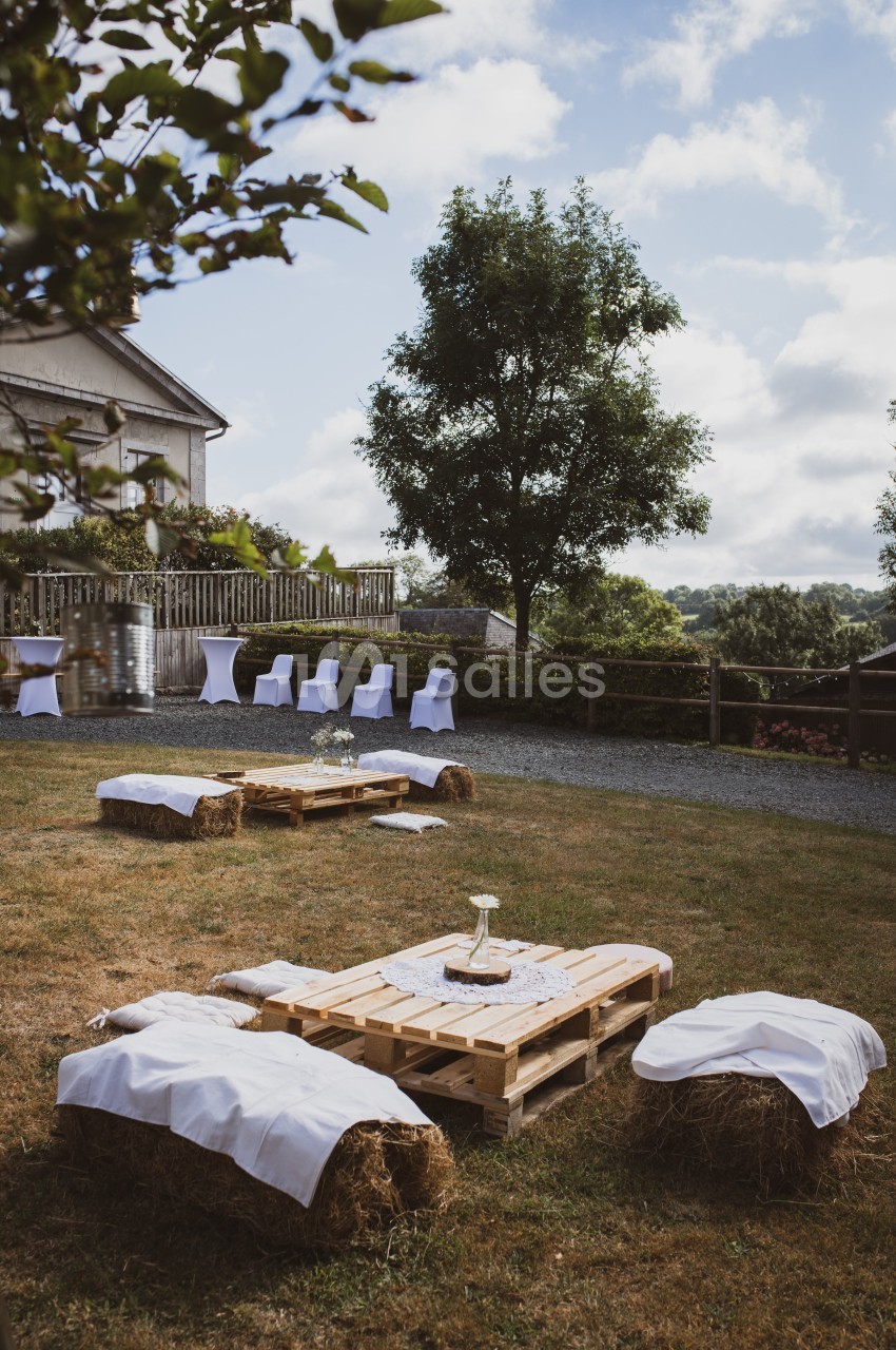 Espace extérieur avec tables en palettes, bottes de paille couvertes de tissu blanc et chaises alignées près d'une maison.