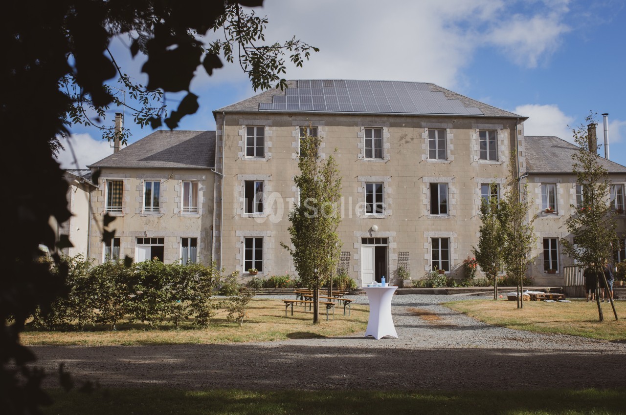 Façade d'un grand bâtiment en pierre entouré d'arbres, avec une table haute blanche au premier plan.