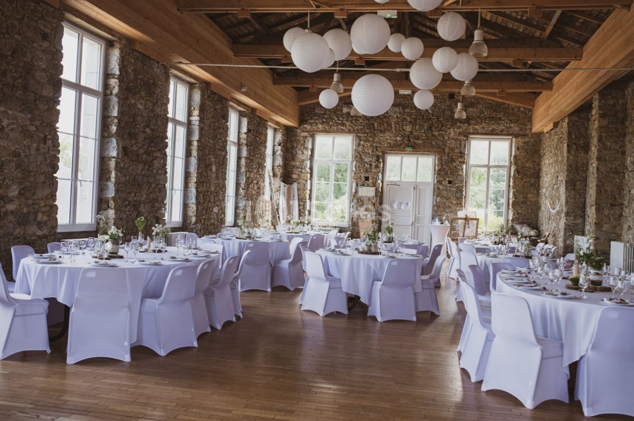 Salle de réception lumineuse avec murs en pierre, tables rondes dressées et décorations suspendues en papier blanc.