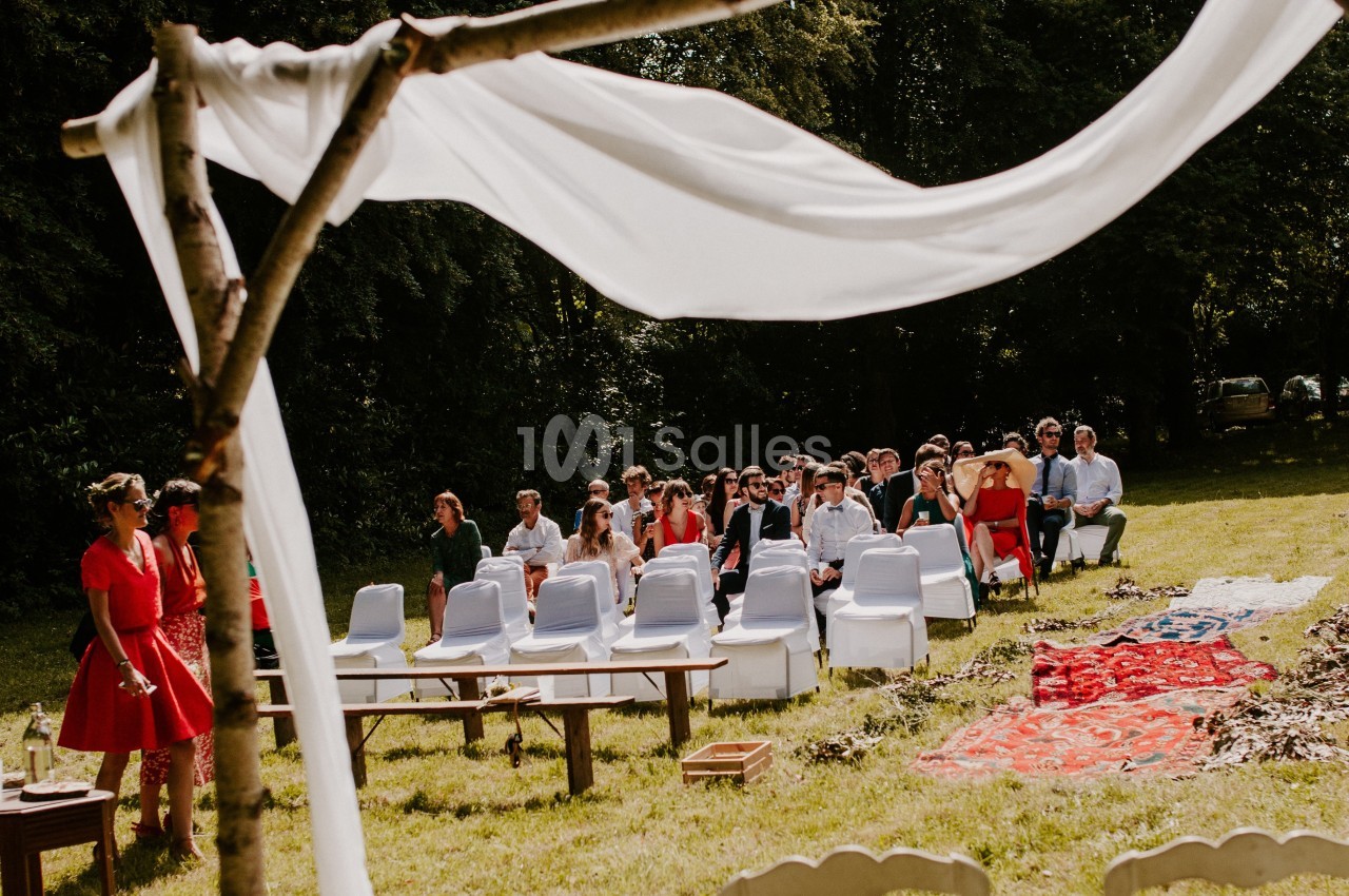 Invités assis en plein air lors d'une cérémonie, sous une arche en bois décorée de tissu blanc, dans un cadre verdoyant.