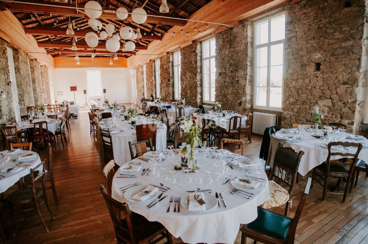 Salle de réception lumineuse avec des tables rondes dressées, entourées de chaises variées, et murs en pierre apparente.