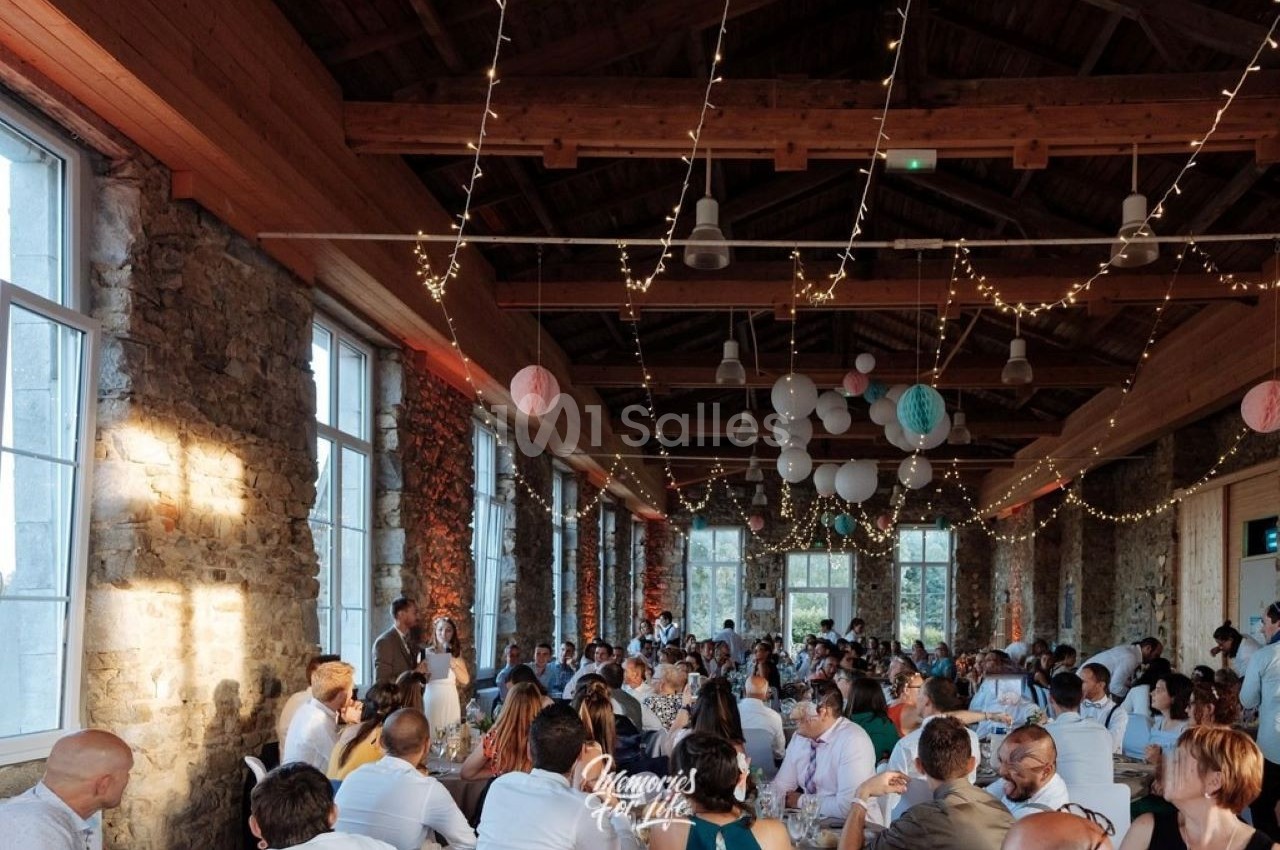 Salle en pierre avec de grandes fenêtres, décorée de guirlandes lumineuses, accueillant un dîner avec de nombreux convives.