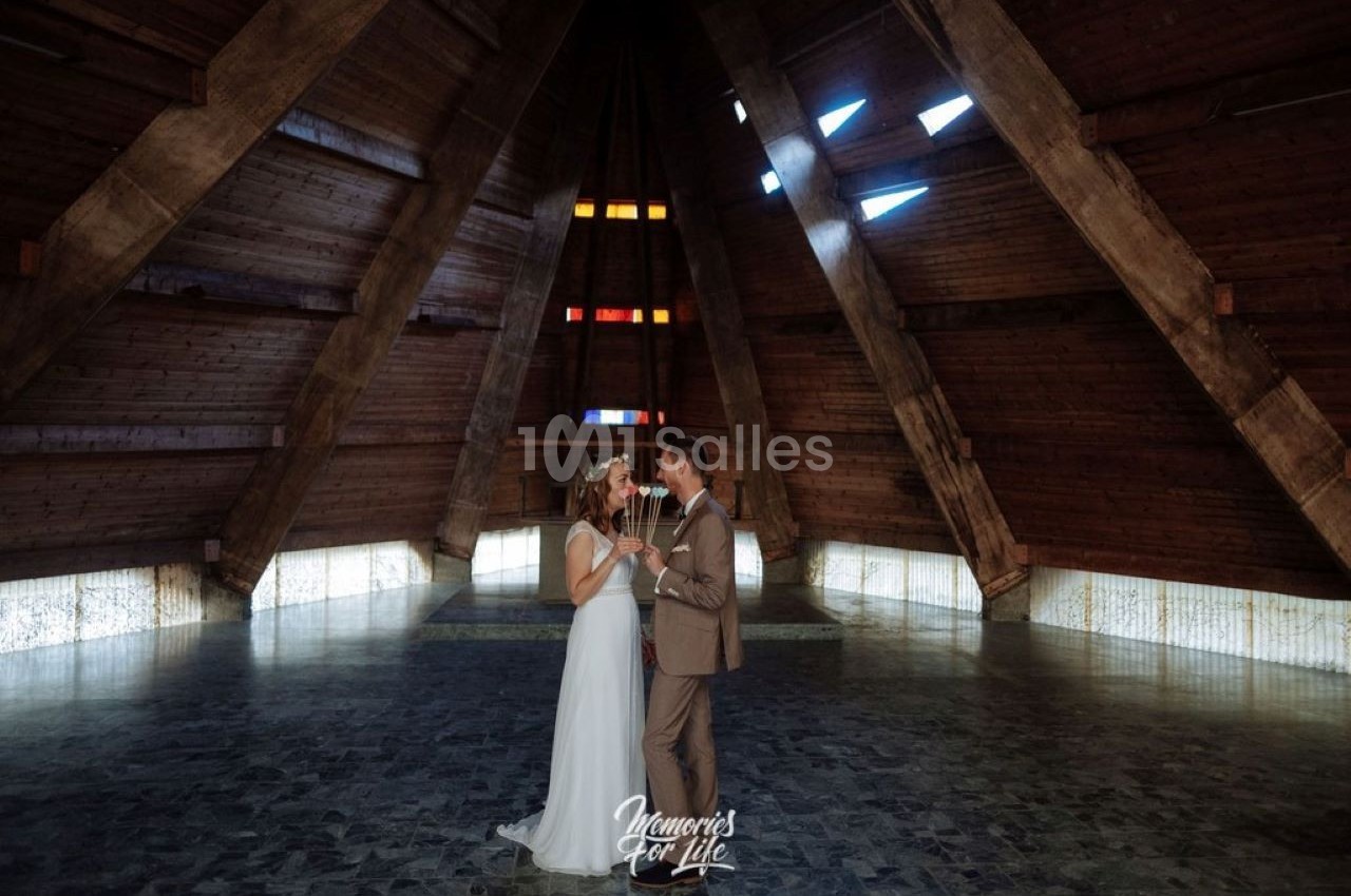 Un couple en tenue de mariage danse dans une salle en bois avec des vitraux colorés et un sol en pierre.