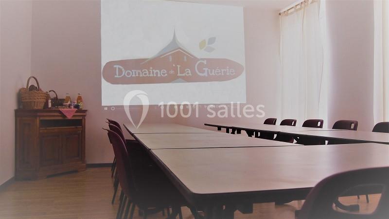Salle de réunion lumineuse avec tables alignées, chaises et projection du logo ’Domaine de la Guérie’ sur un mur.
