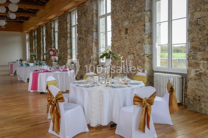 Salle de réception avec tables décorées pour un événement, nappes blanches, chaises ornées de nœuds dorés et grandes…