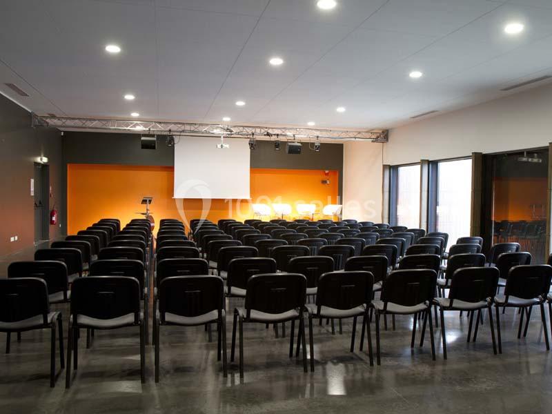 Salle de conférence vide avec des rangées de chaises noires, un écran blanc et un mur orange en arrière-plan.
