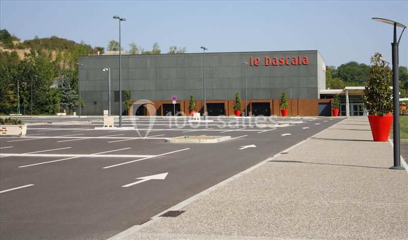 Parking vide devant un bâtiment moderne gris avec l'enseigne ’Le Dastala’ et des arbres en arrière-plan.