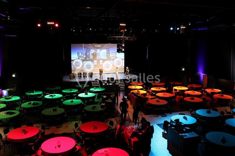 Salle de conférence avec des tables rondes éclairées en vert et rouge, et une scène avec écran projeté en arrière-plan.