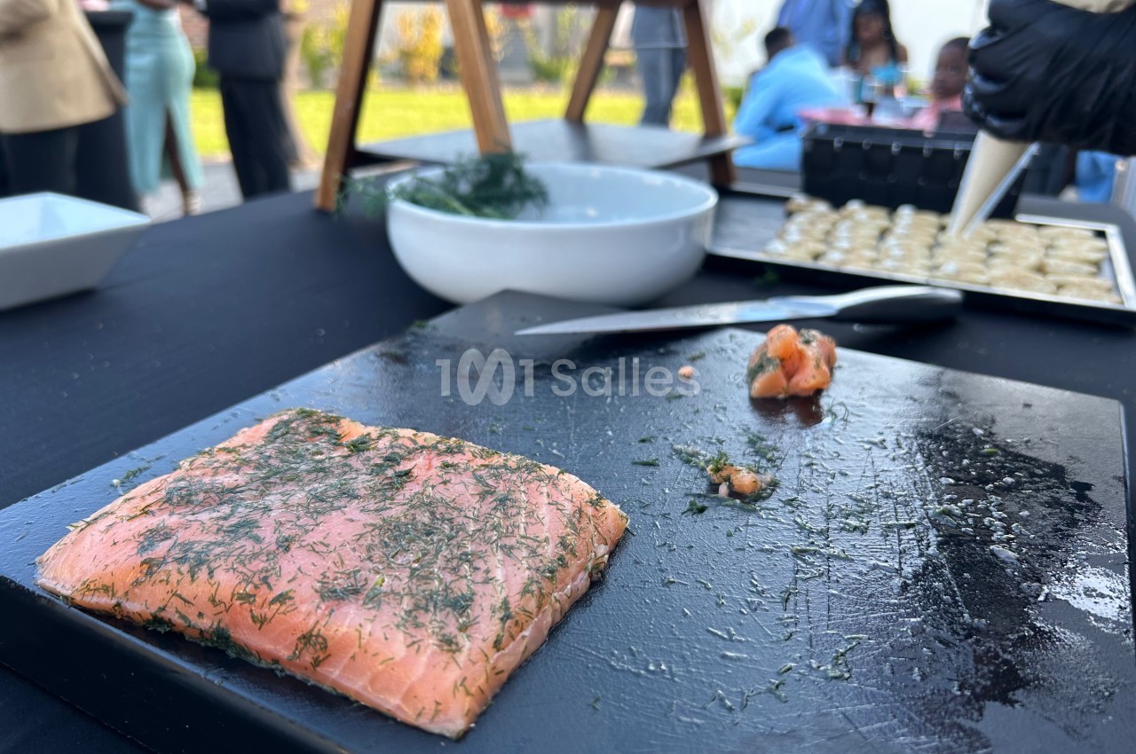 Saumon mariné aux herbes sur une planche noire, avec un bol et des préparations culinaires en arrière-plan.