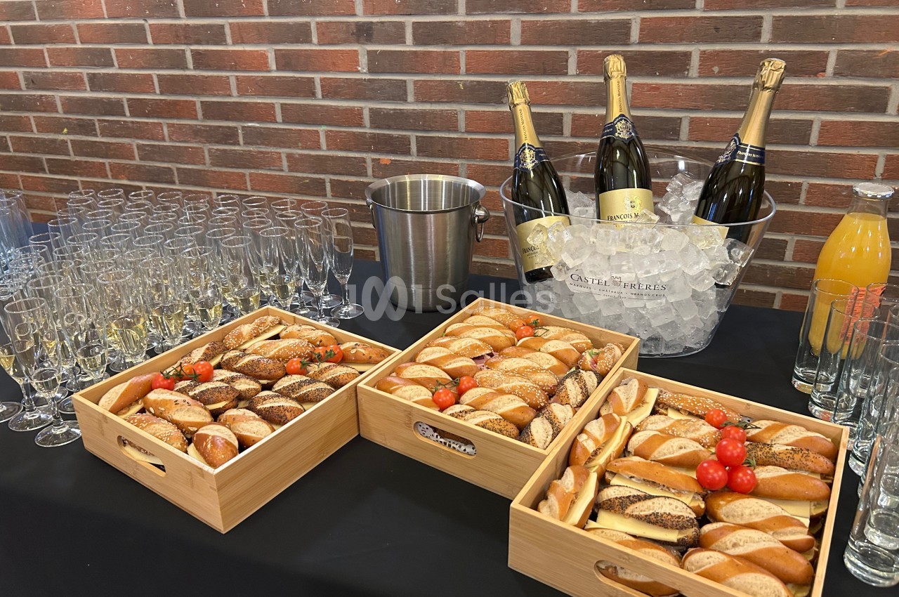 Plateau de pains variés avec tomates cerises, flûtes à champagne, bouteilles sur glace et jus d'orange sur une table.