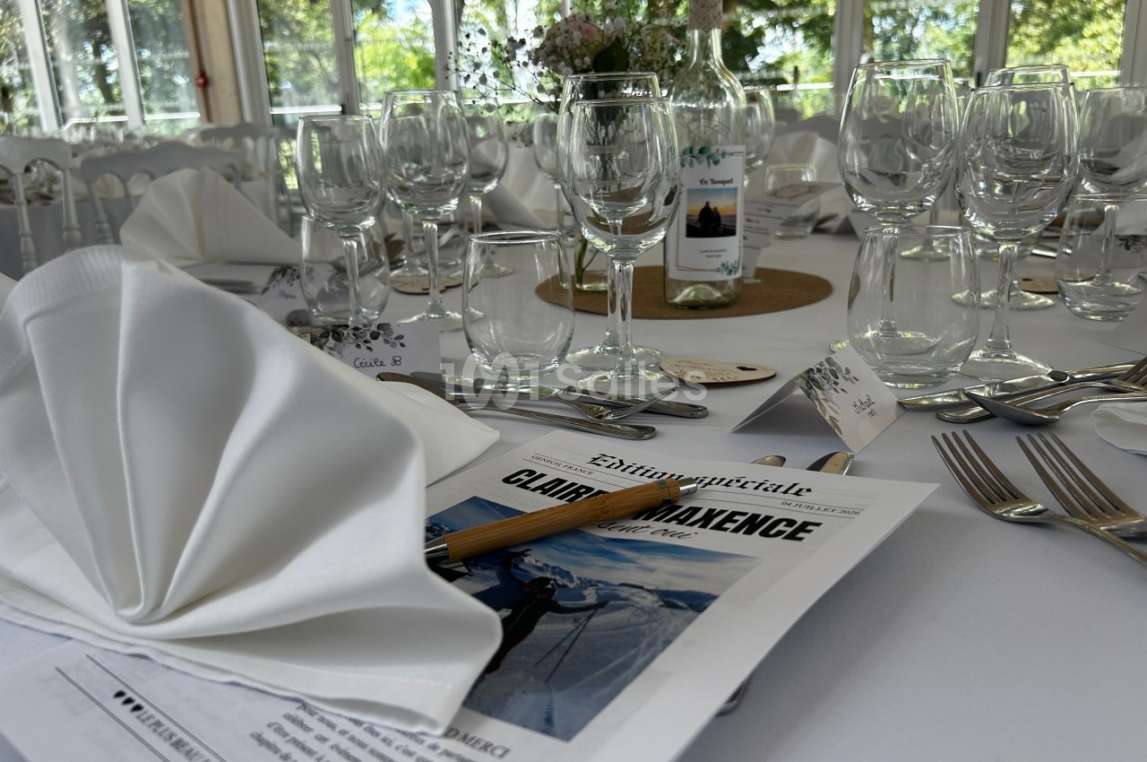 Table dressée pour un événement avec nappes blanches, verres, couverts, menus personnalisés et une bouteille de vin.