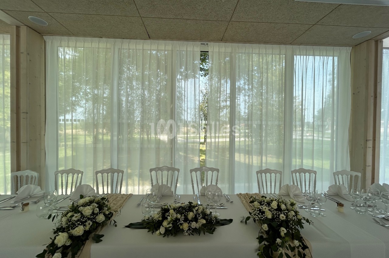 Table décorée pour un événement, ornée de fleurs blanches et de verdure, devant de grandes fenêtres avec rideaux…
