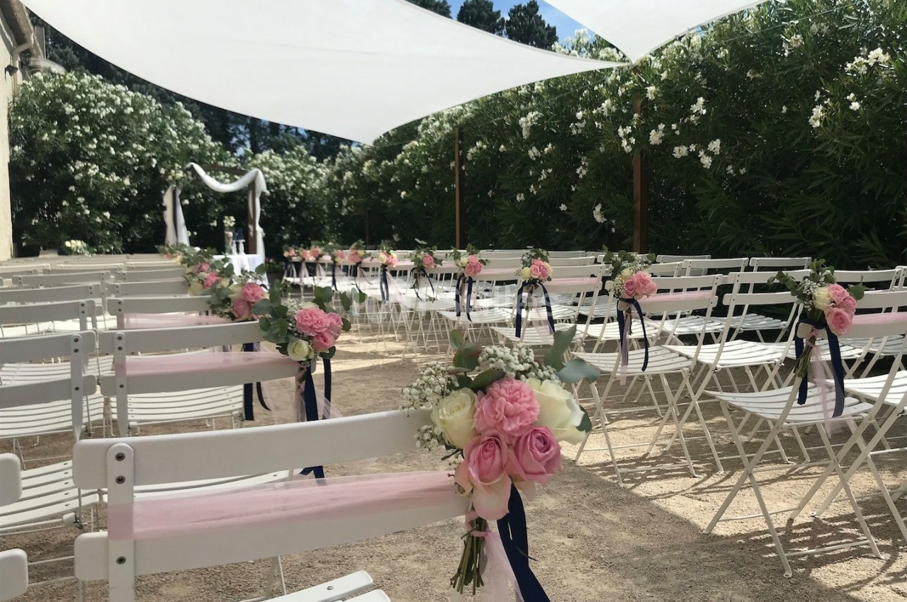 Chaises blanches décorées de fleurs roses et rubans, alignées pour une cérémonie en extérieur sous des voiles d'ombrage.