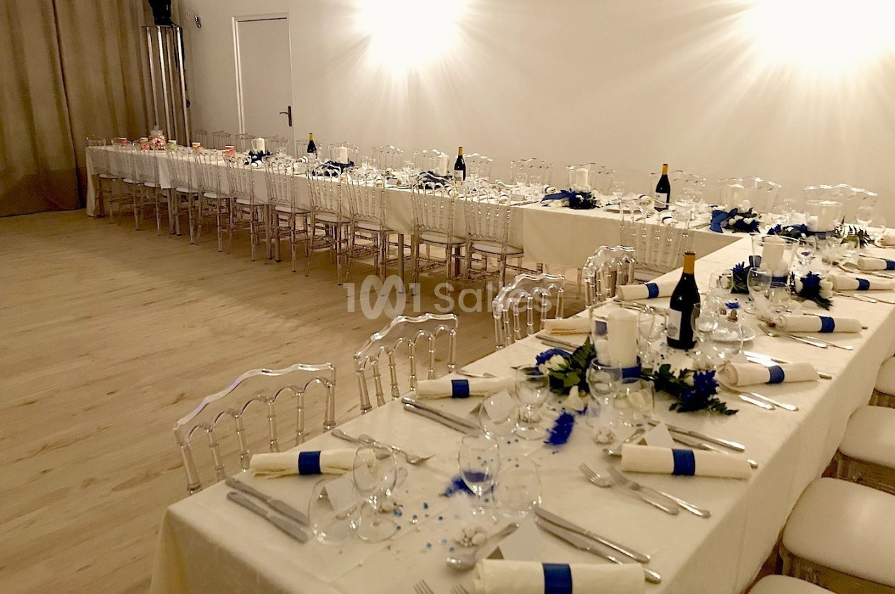Tables élégamment dressées avec nappes blanches, couverts, verres et décorations bleues dans une grande salle lumineuse.