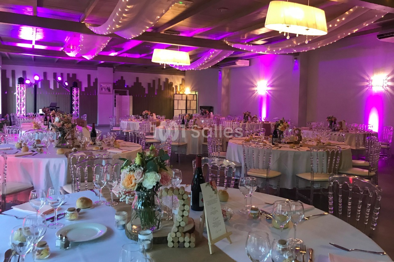 Salle de réception décorée pour un événement, avec tables dressées, chaises transparentes et éclairage violet.