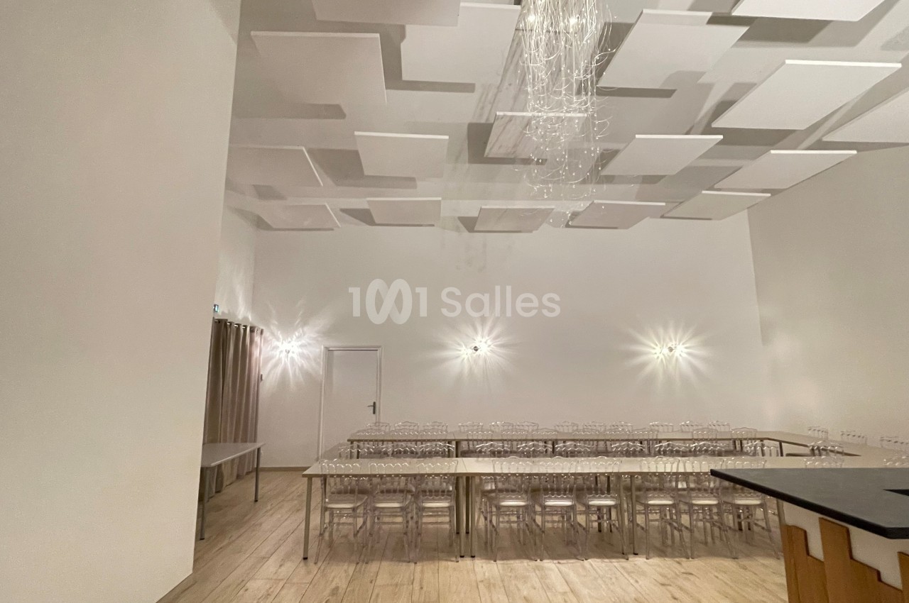 Salle lumineuse avec plafond décoratif, tables alignées avec chaises transparentes et éclairage mural discret.